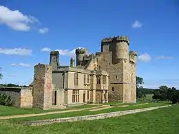 A large stone built ruined building. The nearer part is two stories with square windows; behind is a tall square keep with turrets and battlements. In the foreground is grass with a low stone wall; in the background a blue sky with a few white clouds.