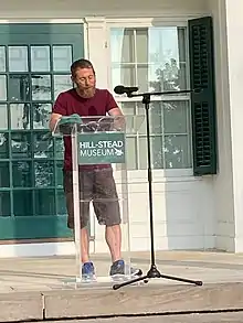 A color, digital full length image of Benjamin S. Grossberg, a man with short brown hair and full beard, wearing glasses, a deep red t-shirt, and dark green shorts standing at a lectern outside on the patio of the Hill-Stead Museum in Farmington, Connecticut.