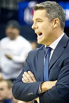 A middle-aged Caucasian man is standing on the court of a basketball arena and shouting toward the left of the frame, with a wedding ring clearly visible on his left hand.