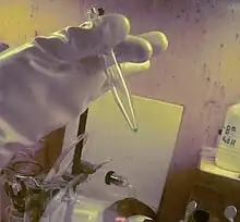 A very small sample of a blue liquid in a plastic pipette held by a hand wearing heavy protection equipment