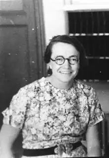 A smiling woman with glasses in a patterned dress with a dark belt sitting in front of a wood door and some shelving.