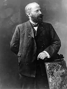 Eduard Bernstein standing next to a chair and looking rightwards. He is resting his hand on the chair.