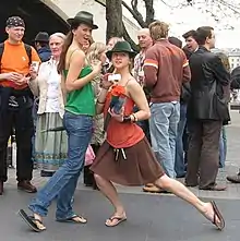 Two women with black hats are standing in front of a crowd of people, and are posing for cameras. The left woman is wearing a green shirt and jeans. The right woman has a red shirt with a brown skirt.