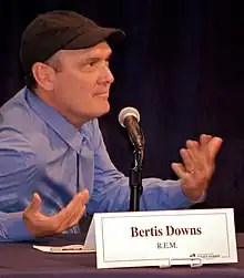 Bertis Downs looking to the camera's left and gesturing with his hands as he sits at a table behind a placard with his name