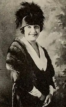 A young woman standing, smiling, wearing a hat with a large dark plum and a fur stole; her dress has a large white collar and cuffs.