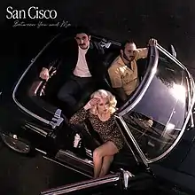 Two men and one woman sit in a retro car, looking up at the camera