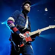 A man wearing a dark cap and a jacket with colourful details plays the guitar