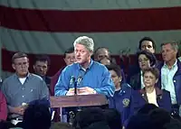 Bill Clinton is standing at a podium speaking to a crowd. The former mayor of Grand Forks is at the right of the image.