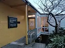 The entrance of the Bill Manhire House, a seemingly small building, with a yellow wall and tree's around.