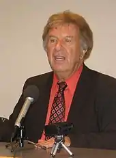 A man, sitting, speaking in front of a microphone