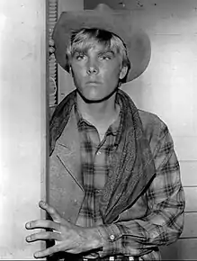 A black-and-white photo of a young Caucasian man with light-colored hair. He is wearing a cowboy hat and flannel shirt. He looks to the left of the camera, holding his left hand against the wall.