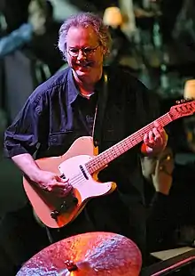 A man wearing glasses, playing a guitar and standing behind a cymbal.