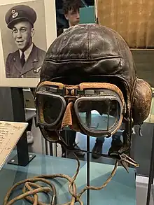 A photograph of a museum display containing items once belonging to Billy Strachan. On this display there are flight goggles, a flight helmet, and a photograph of Billy Strachan