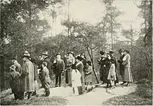Sepia-tone image of men in suits and women in long dresses and hats, facing left, looking at something out of frame