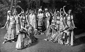  A group of female dancers in flowing robes strike exaggerated poses around a central group of two men and a woman.