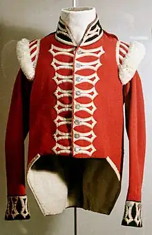 A color photograph of a red coat cut high-waisted in the front and slightly longer in the back so that it reaches the hips. There is white cording in a spade-like design along the fasteners and wool puffs along the shoulders. The collar is high and black but opens at in a V-shape along the neck. The cuffs are also black with white cording in the same spade-like design like that along the front fasteners.