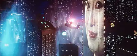 Screenshot of a police spinner flying through a cityscape next to a large building which has a huge face projected onto it. In the distance a screen can be seen with writing and pictures on it