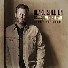 American singer Blake Shelton stands in front of a light brown backdrop displaying the "Blake Shelton Featuring Gwen Stefani, Happy Anywhere"