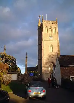 Square three stage stone tower. To the right is a building with a white wall and in the foreground a parked car.