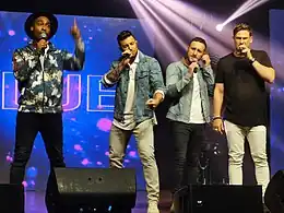 An image of 4 male performers onstage singing to a hand held microphone