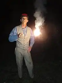 A man is standing in the dark. He is holding out a short stick at mid-chest level. The end of the stick is alight, burning very brightly, and emitting smoke.