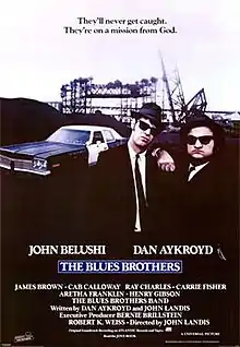 Movie poster with two of the main characters on the right side of the image: They are both wearing black suits, hats, and sunglasses and facing forward. The man on the right is resting his arm on the shoulder of the man on the left. A police car is present on the left side of the image behind them. At the top of the image is the tagline, "They'll never get caught. They're on a mission from God." At the bottom of the poster is the title of the film, cast names, and production credits.