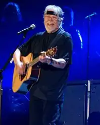 A grey-haired man wearing dark clothing and a headband, playing a guitar