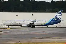A white plane with a navy blue eskimo on the tail, the word "Alaska" painted in silver, a logo of Boeing painted in silver in the middle, and the words "100 years strong" painted in silver across the fuselage, taxis at an airport taxiway