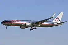 Side quarter view of twin-engine jetliner in flight, with extended gear; bare metal with red, white, and blue cheatlines, as well as two "A"s and a stylized eagle on the tail