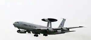 Four-engined jet aircraft in-flight with landing gear partially extended. A large disc-shaped radar perches on two convergent struts on the aft fuselage.