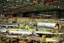 Airplane assembly hall, featuring heavy machinery. Large cylindrical airplane sections and wings are readied for mating with other major components. Above are the cranes which ferry heavy and outsize parts of the 747.