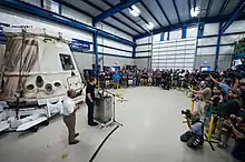 Musk is pictrued delivering a speech to SpaceX employees while standing at a podium besides a capsule in 2012