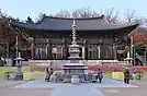 A head-on shot of the Bongeunsa buddhist temple. The temple has a traditional Korean roof and is adorned with various decorative designs and hanja characters. In front of the temple are a few people surrounding a large stone pillar.
