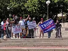 A group of people hold signs and flags, including a Trump 2020 flag, while standing against police barricades around Boston's Government Center.