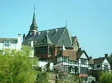 A church in an elevated position with a steep roof and a pinnacle to the left. In front of the church are black-and-white buildings and in the left foreground are the branches of a tree