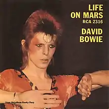 A photo of a red-haired man wearing red face make-up, wearing a red V-neck shirt with his arms out against a black backdrop. The yellow-lettered words "Life on Mars" appear in the top left corner, with "RCA 2316" under it and "David Bowie" under that.