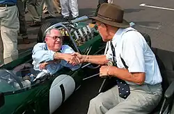 Two elderly men shake hands on a race starting grid. One is sitting in a green racing car, the other in a wheelchair.