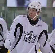 Head and upper body of a hockey player in white uniform with a picture of a crown in the middle. He smiles slightly, looking forward and to his right.