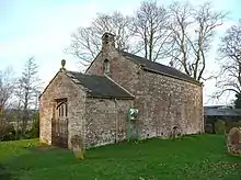A small bare stone building seen from an angle, a lower section with a doorway at the front, and a larger, higher section with a bellcote at the back