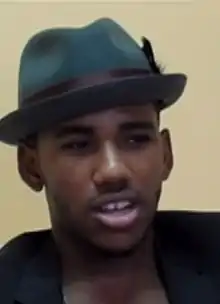 A head shot of a young man wearing a dark fedora hat and a blazer jacket. He is talking at a press event.