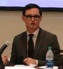 Frye in a dark suit sitting and speaking on a panel