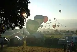 A large number of hot air balloons taking off from a field which is surrounded by tents and stalls. The sun is low in the sky and balloons can be seen flying into the distance.