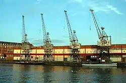 A long two-storey building with 4 cranes in front on the quayside. Two tugboats are moored at the quay.