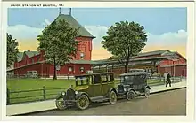 A color postcard of a train station with two early automobiles