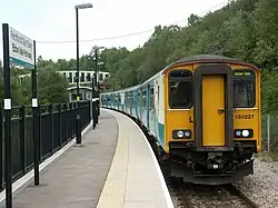 Four carriage train, on single curved track, at station. Steep wooded bank on one side of the track. Platform to the other. Station sign, in Welsh above English, in the foreground. Modern bridge over the track in the background.