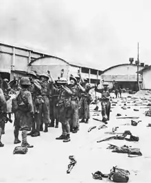 Many British soldiers hold their hands in the air, while Japanese soldiers in the foreground level their weapons at them. Weapons and kitbags litter the ground on the right side of the photo.