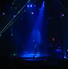 Faraway image of a female performer. She is walking through a water screen that is falling from the ceiling. A blue spotlight is on her. She is wearing blue jeans and a plastic cowboy hat. Green laser lights surround the stage.