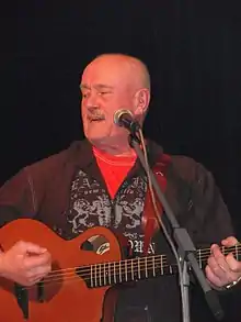 bald, middle-aged white male playing a guitar in front of a microphone, looking right of camera