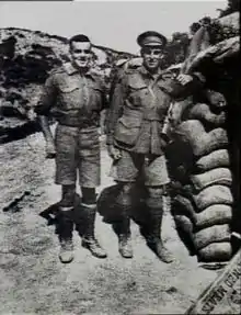 Two men in military uniform standing side-by-side. The man on the left is slightly shorter and stockier, and the man on the right has his arm leaning on a stacked up pile of sandbags.