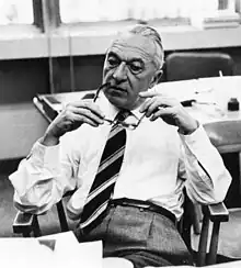 An old man sitting in a chair and wearing a shirt with a tie. He is holding his glasses with both hands and is sitting in front of his desk